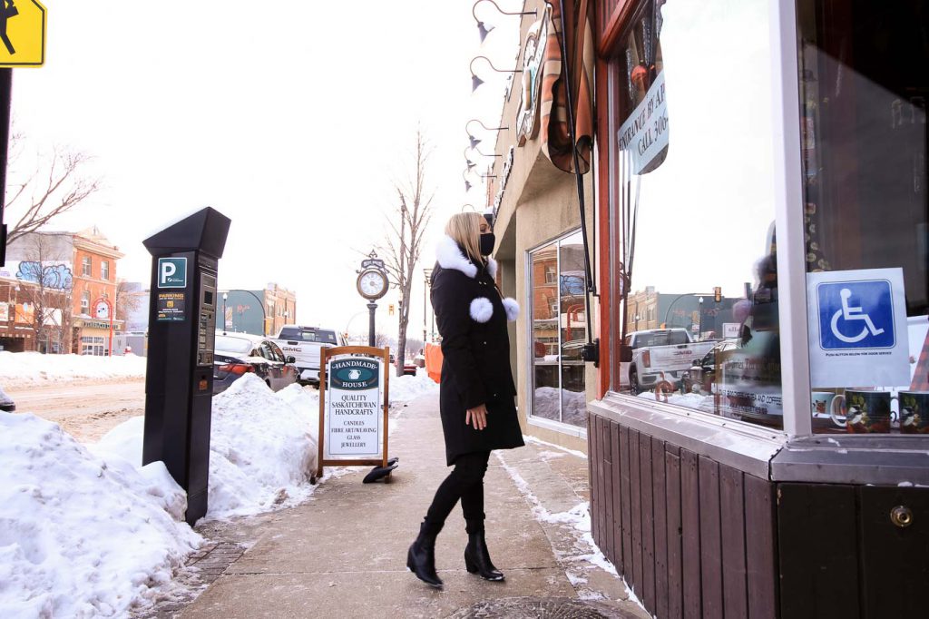 A blonde woman with a black mask on, wearing a black ski jacket with a white hood and pom poms, looks into the window of a building on Broadway Avenue in Saskatoon. It's winter and snow lines the street.