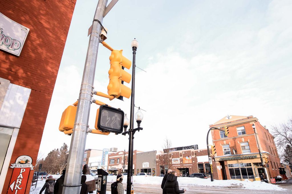 People wait at a crosswalk on Broadway Avenue in Saskatoon. You can see Starbucks Coffee across the street. It's winter and there are piles of snow.